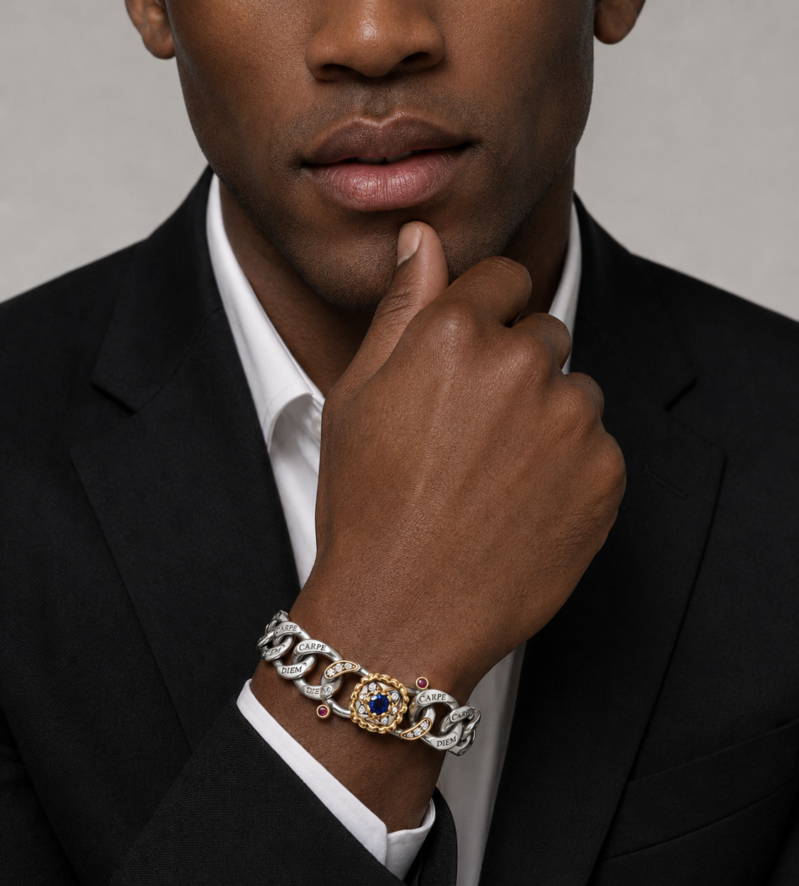 A close-up of a man in a black suit and white shirt wearing a luxury chain-link bracelet on his wrist. The bracelet features interlocking silver and gold links, with one central gold link detailed with a blue gemstone and small clear stones.