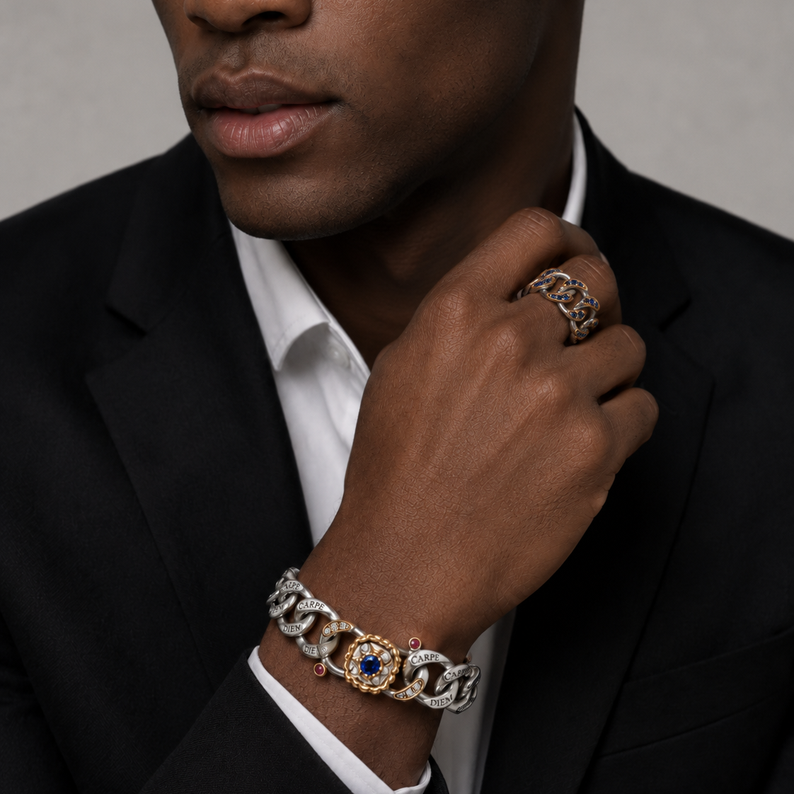 A close-up of a man wearing a black suit jacket and white dress shirt, showcasing a gold and silver chain bracelet with a central blue gemstone on his wrist, along with a matching chain ring on his finger.