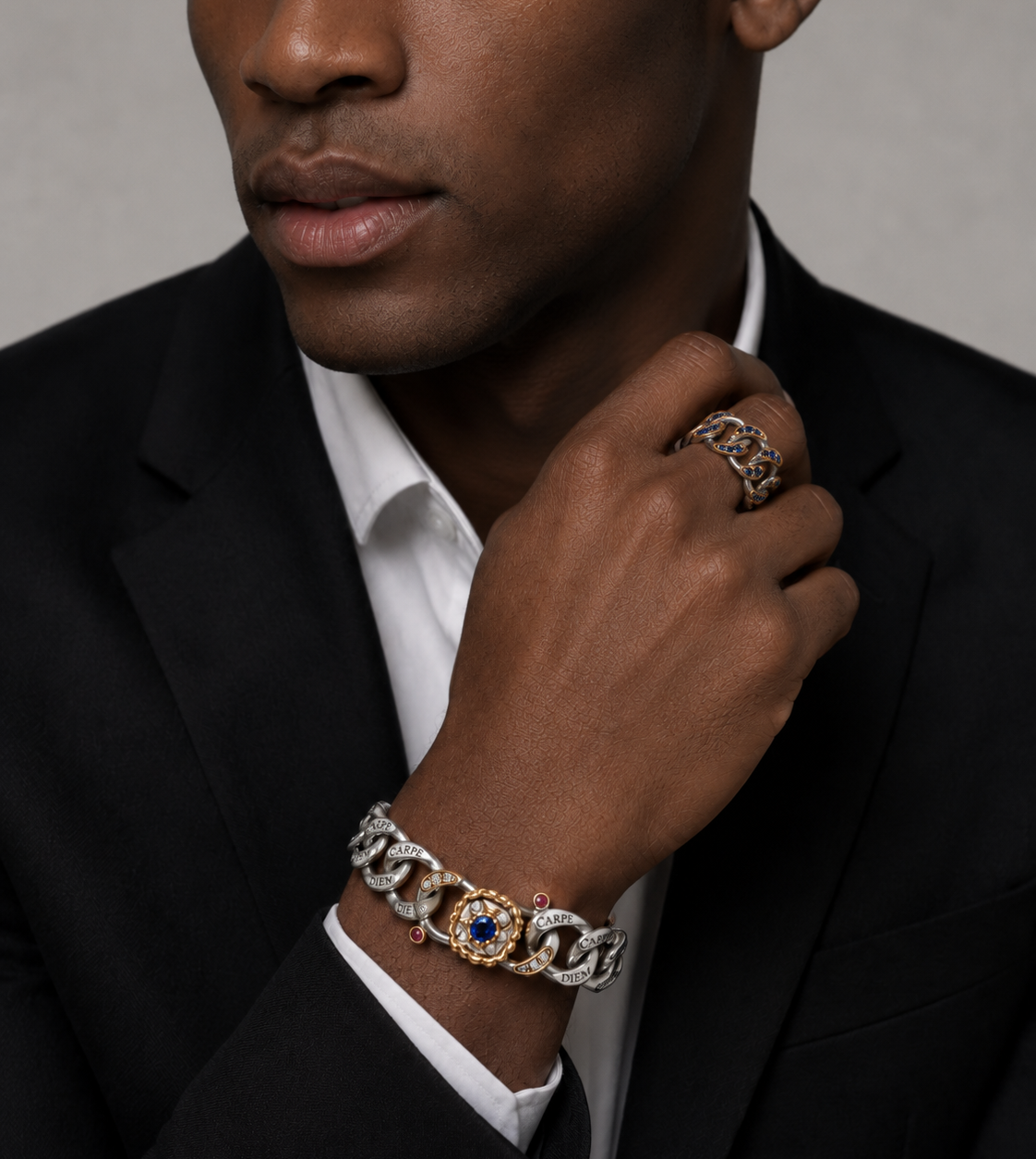 A close-up of a man wearing a black suit jacket and white dress shirt, showcasing a gold and silver chain bracelet with a central blue gemstone on his wrist, along with a matching chain ring on his finger.