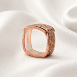 A rose gold ring with an ornate, engraved patterned band, resting on a draped white fabric background.